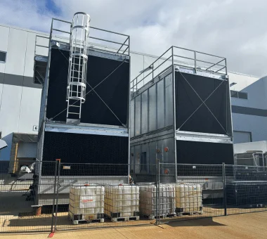 Two large industrial cooling units with protective railings and a ladder are positioned outside a building.