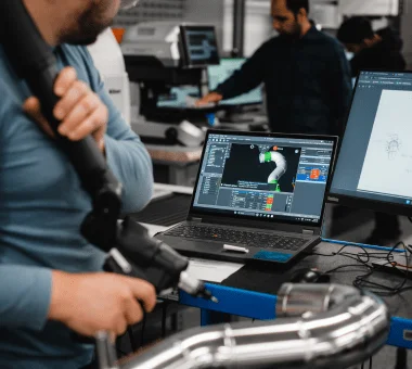 A technician operates a handheld device, inspecting a metal component while viewing data on a laptop.